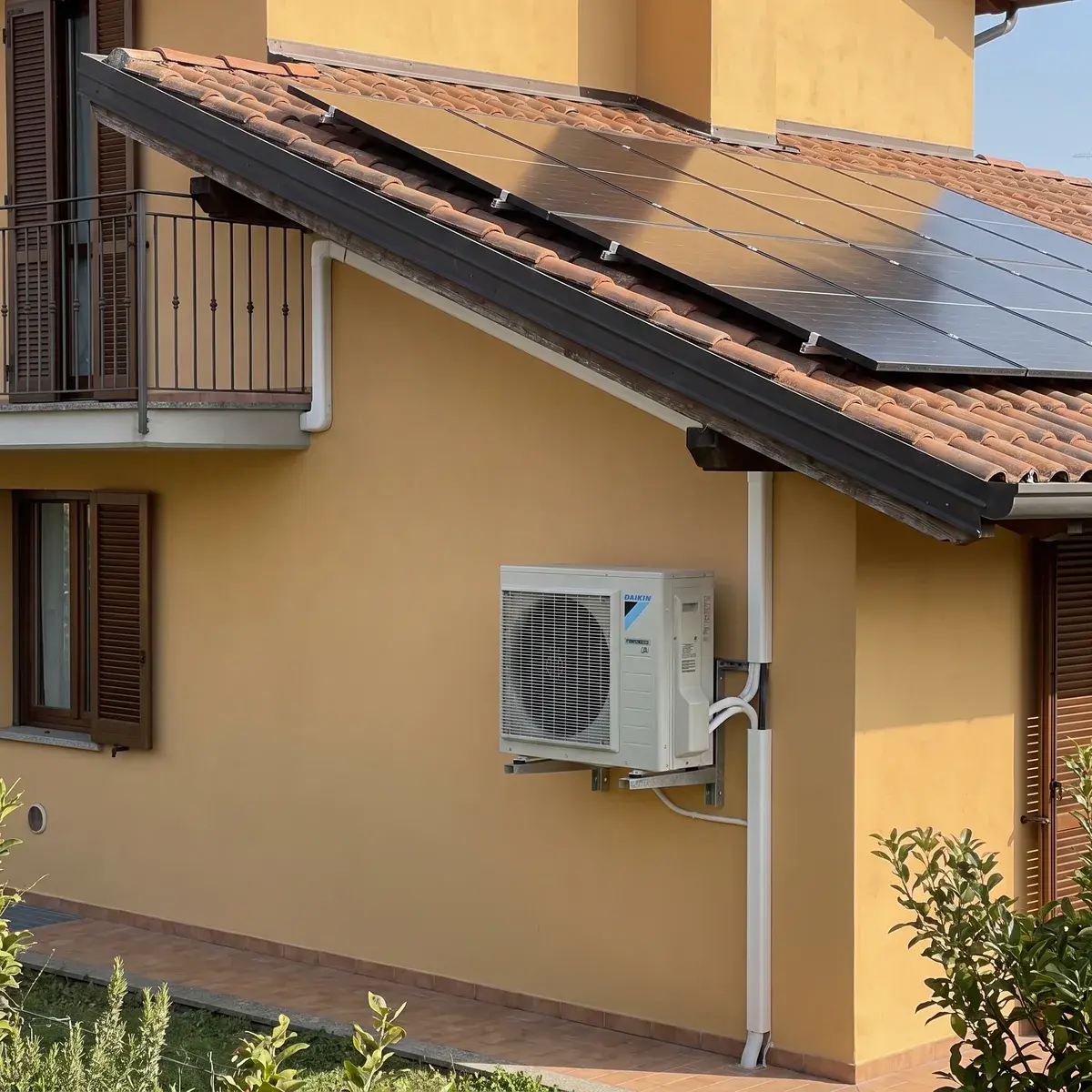 Fianco di una villetta in intonaco ocra con moduli LONGi Hi-MO X10 sul tetto in coppi e unità esterna Daikin Perfera su staffe a parete.