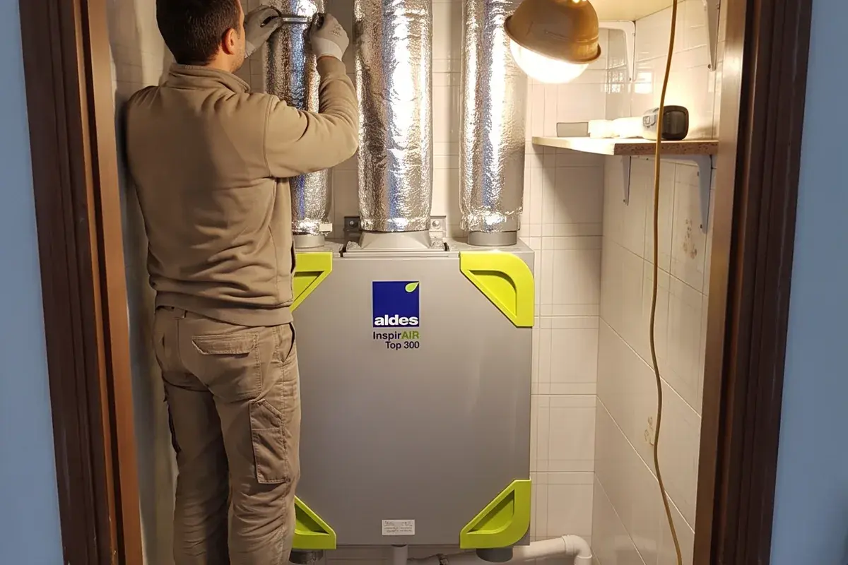 Installatore che stringe una fascetta sui canali isolati sopra un recuperatore centralizzato Aldes InspirAIR Top 300 montato in un ripostiglio piastrellato.