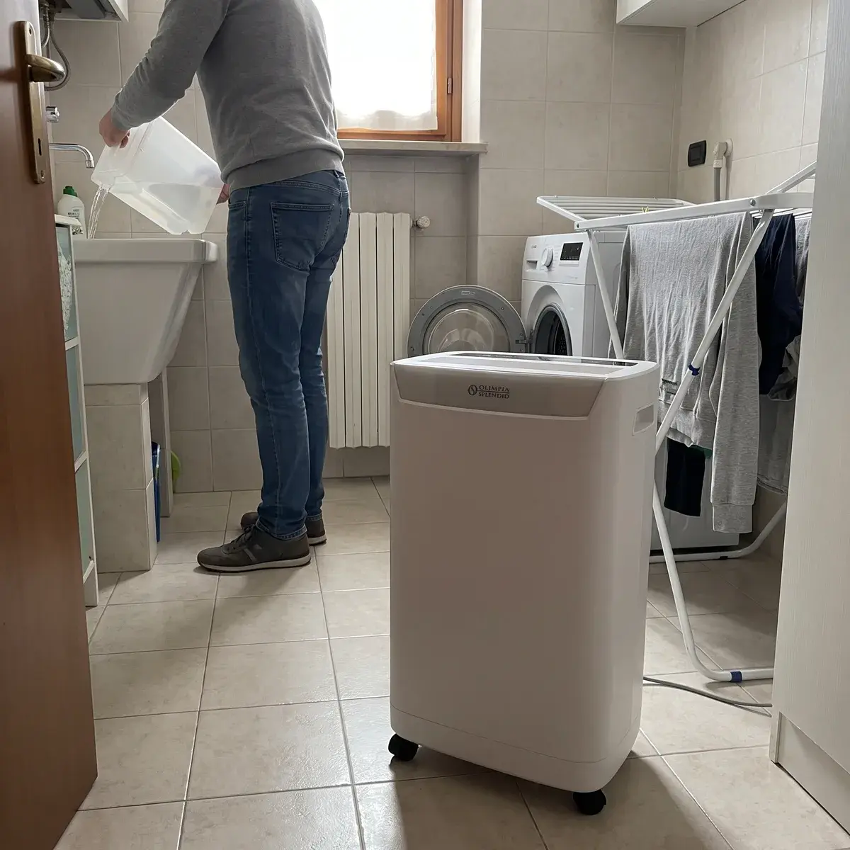 Deumidificatore a compressore Olimpia Splendid Aquaria S1 16 P al centro di una lavanderia con il bucato steso, mentre una persona svuota la tanica nel lavatoio.