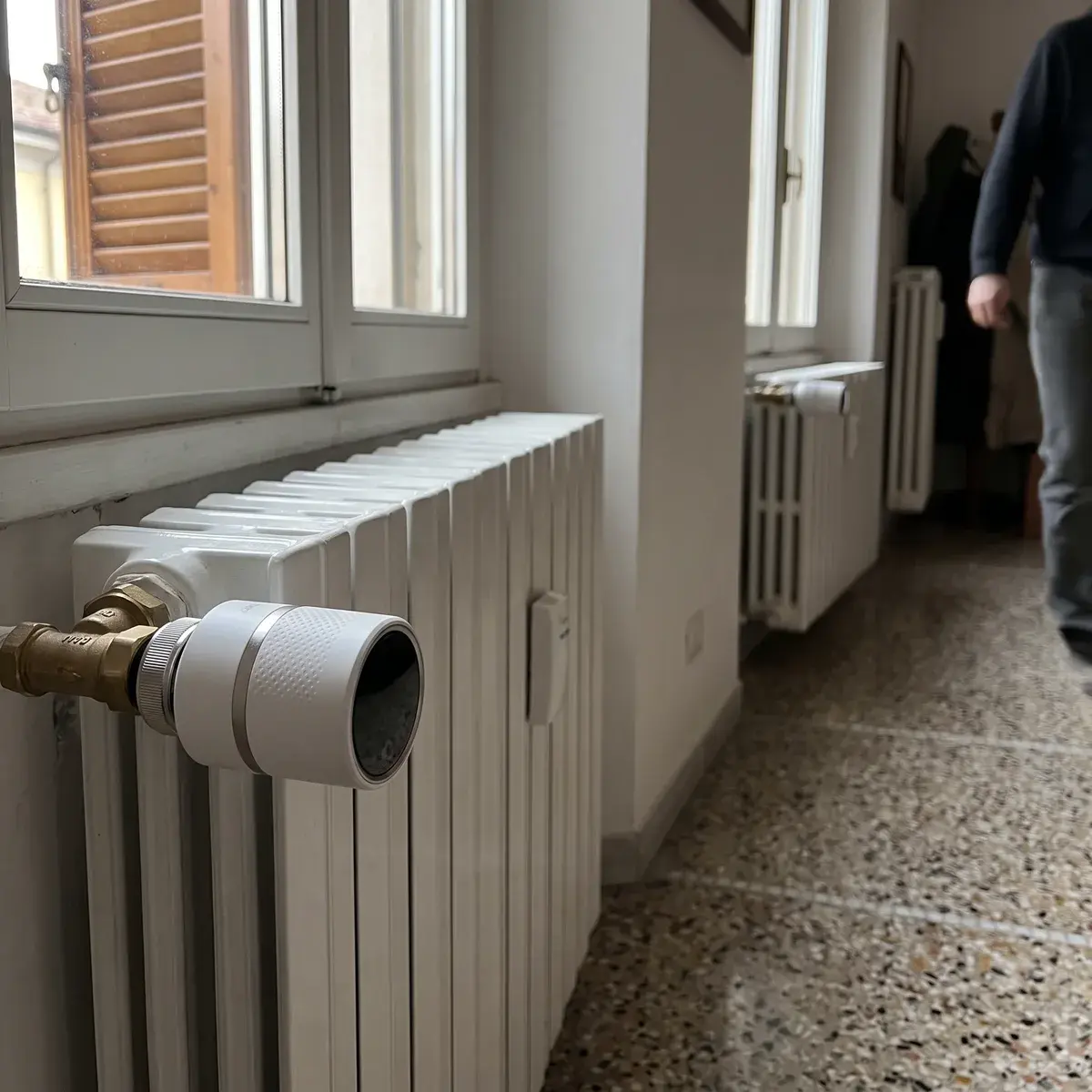Testa termostatica tado X montata sulla valvola in ottone di un radiatore sotto la finestra, con le persiane in legno sullo sfondo e un secondo radiatore più lontano.
