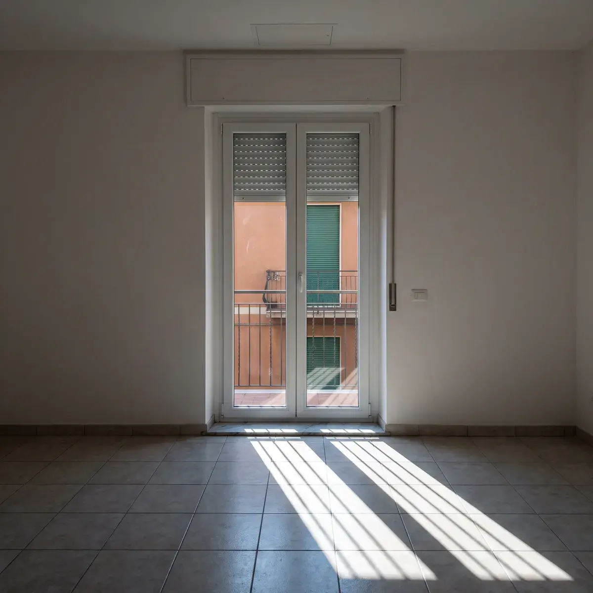 Portafinestra in PVC bianco con la tapparella abbassata a metà e il cassonetto sopra, in una stanza con pavimento in ceramica attraversato da strisce di sole basso; fuori, la ringhiera del balcone e una palazzina con le tapparelle chiuse.