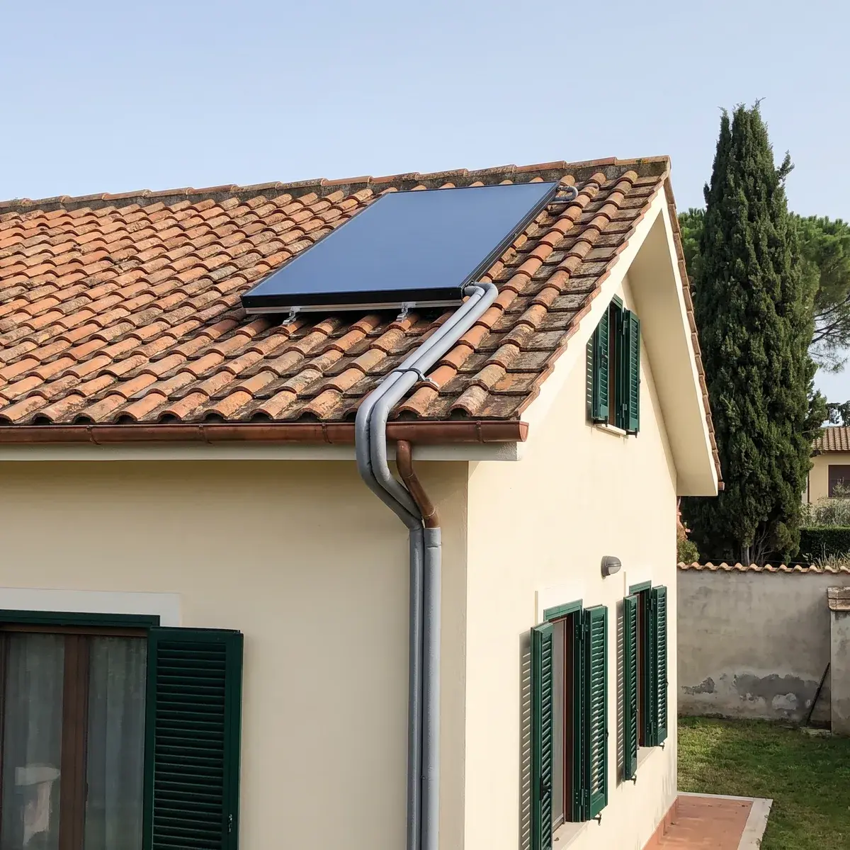 Collettore solare termico piano Bosch Solar 7000 FT 226-2 montato in aderenza sul tetto in coppi di una casa con intonaco chiaro e persiane verdi, con i tubi isolati che scendono lungo il muro e un cipresso sullo sfondo.