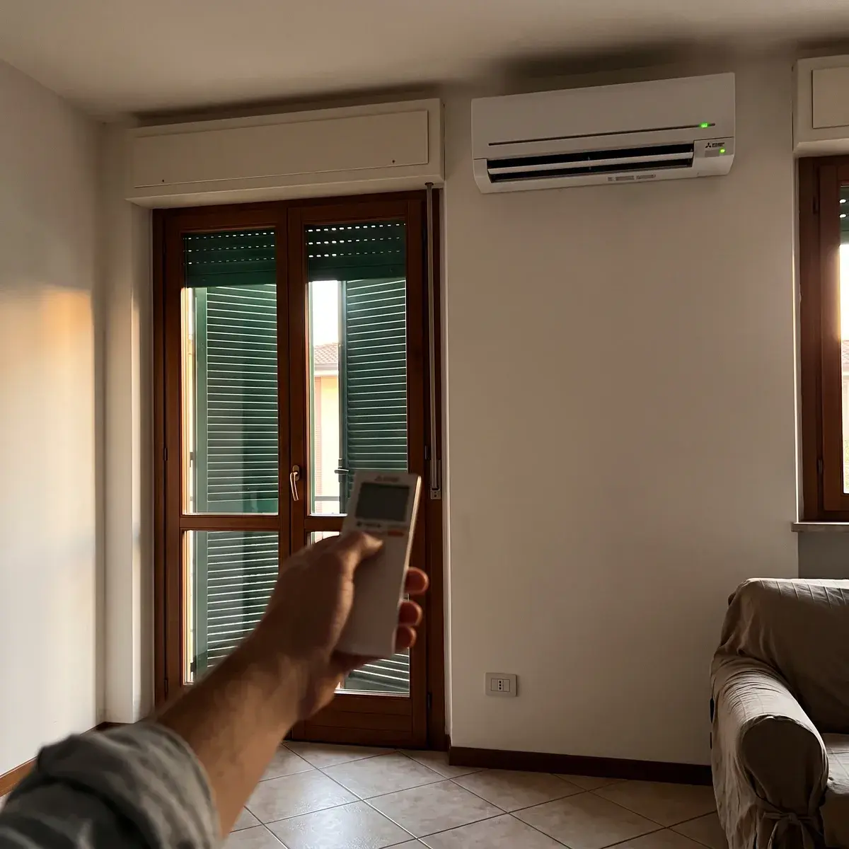 Una mano punta il telecomando verso un climatizzatore Mitsubishi Electric serie MSZ-AP a parete, accanto a una porta-finestra con le tapparelle abbassate, in un soggiorno con il pavimento in piastrelle.