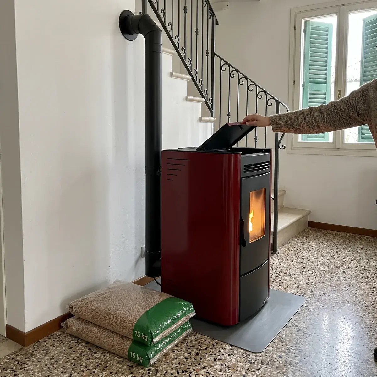 Stufa a pellet ventilata Extraflame Ketty 5.0 bordeaux su una lastra salvapavimento in un soggiorno con pavimento alla veneziana: una mano chiude il coperchio del serbatoio, il tubo dei fumi sale nel muro e accanto ci sono due sacchi di pellet da 15 kg.