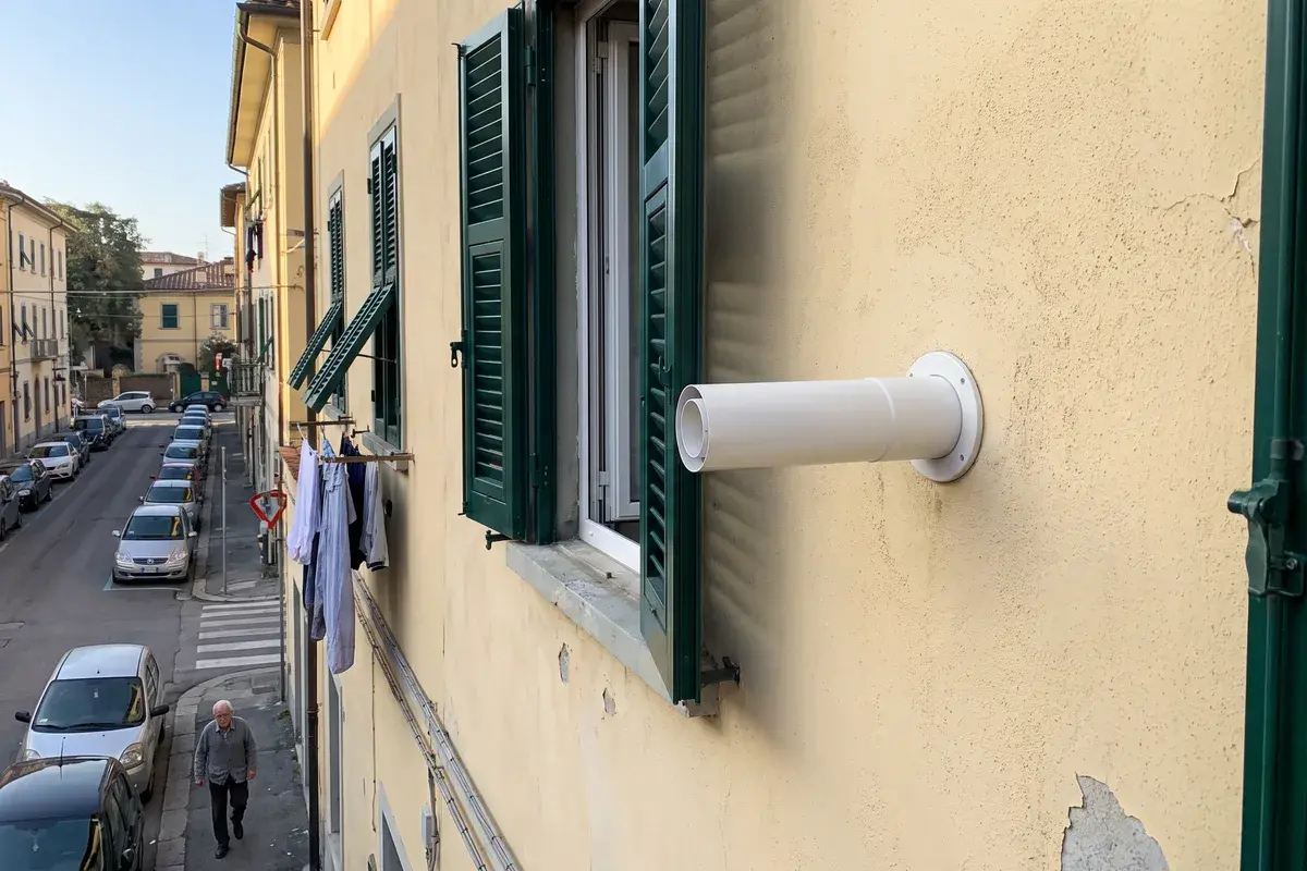 Terminale coassiale bianco di scarico fumi che sporge dalla facciata gialla di un condominio accanto a una finestra con persiane verdi.