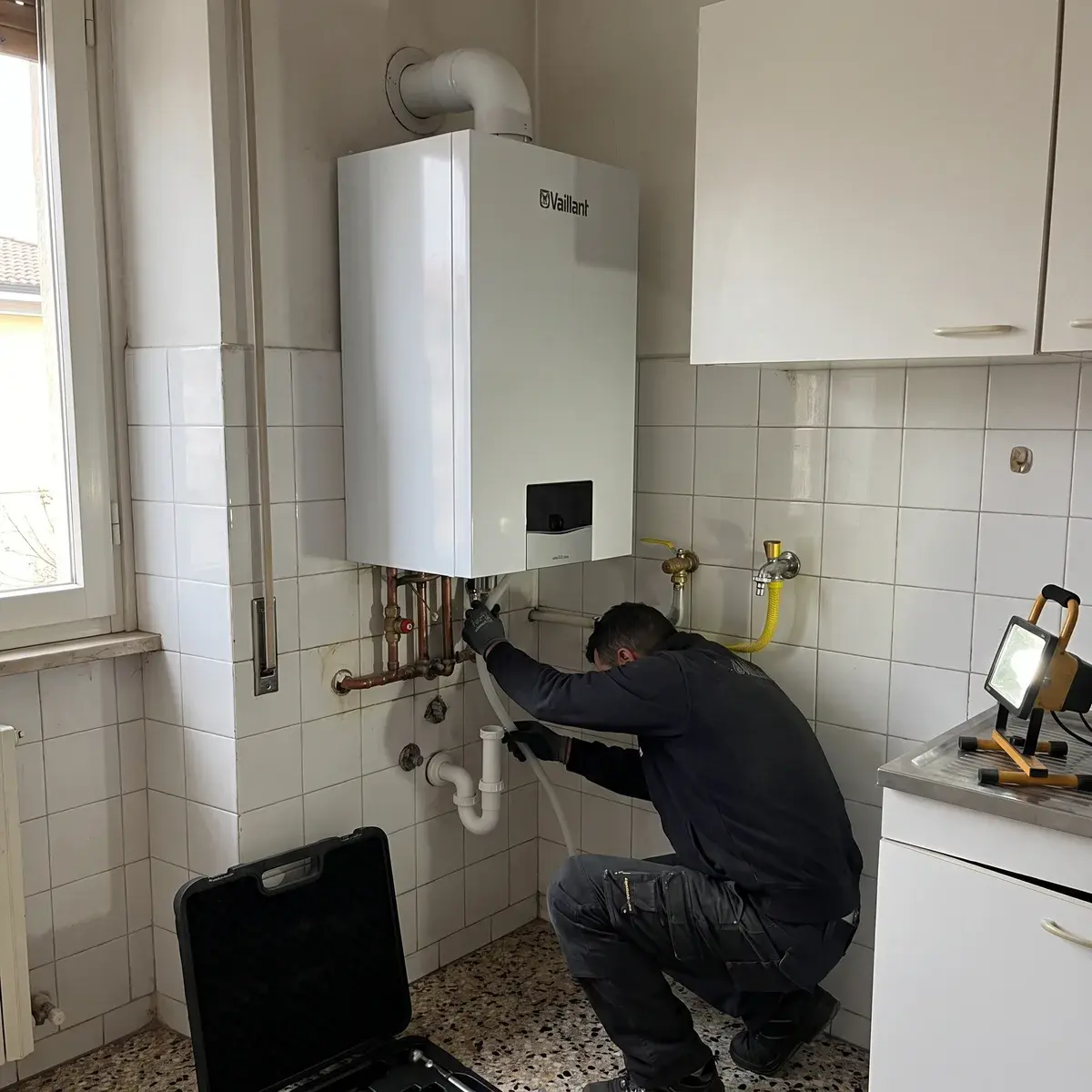 Tecnico accovacciato sotto una caldaia murale a condensazione Vaillant ecoTEC plus che collega il tubo della condensa al sifone di scarico, nella cucina piastrellata di un appartamento.