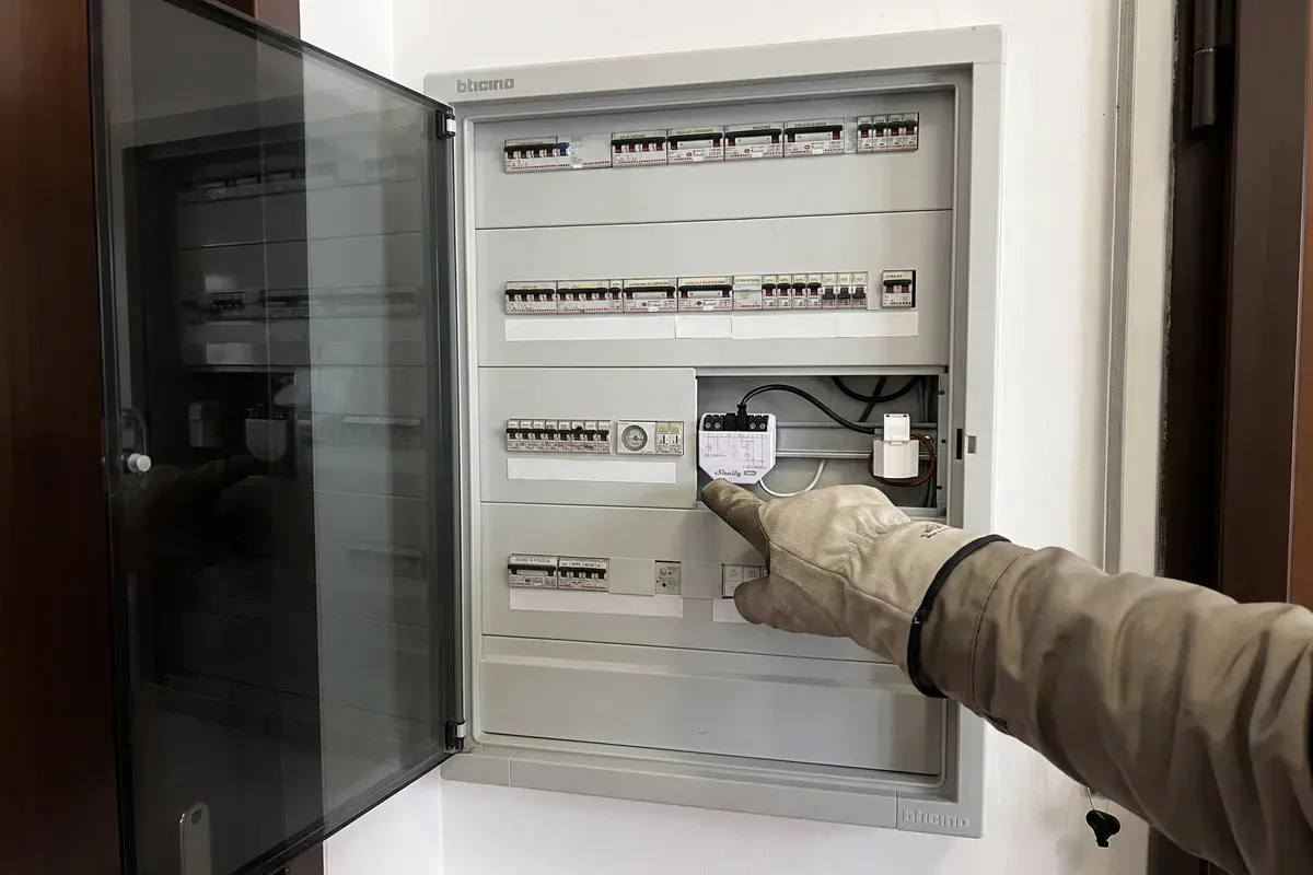 Elettricista con guanti isolanti che indica un misuratore Shelly EM Gen3 montato sulla guida DIN di un quadro elettrico BTicino a incasso con lo sportello fumé aperto.