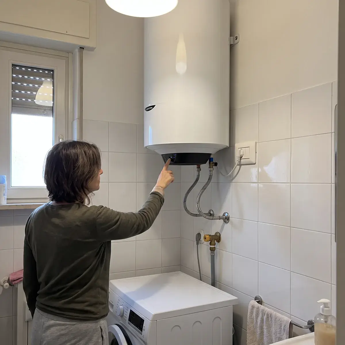Una persona regola il termostato sul pannello di uno scaldabagno elettrico verticale Ariston Pro1 Eco 80 appeso sopra la lavatrice in un bagno.