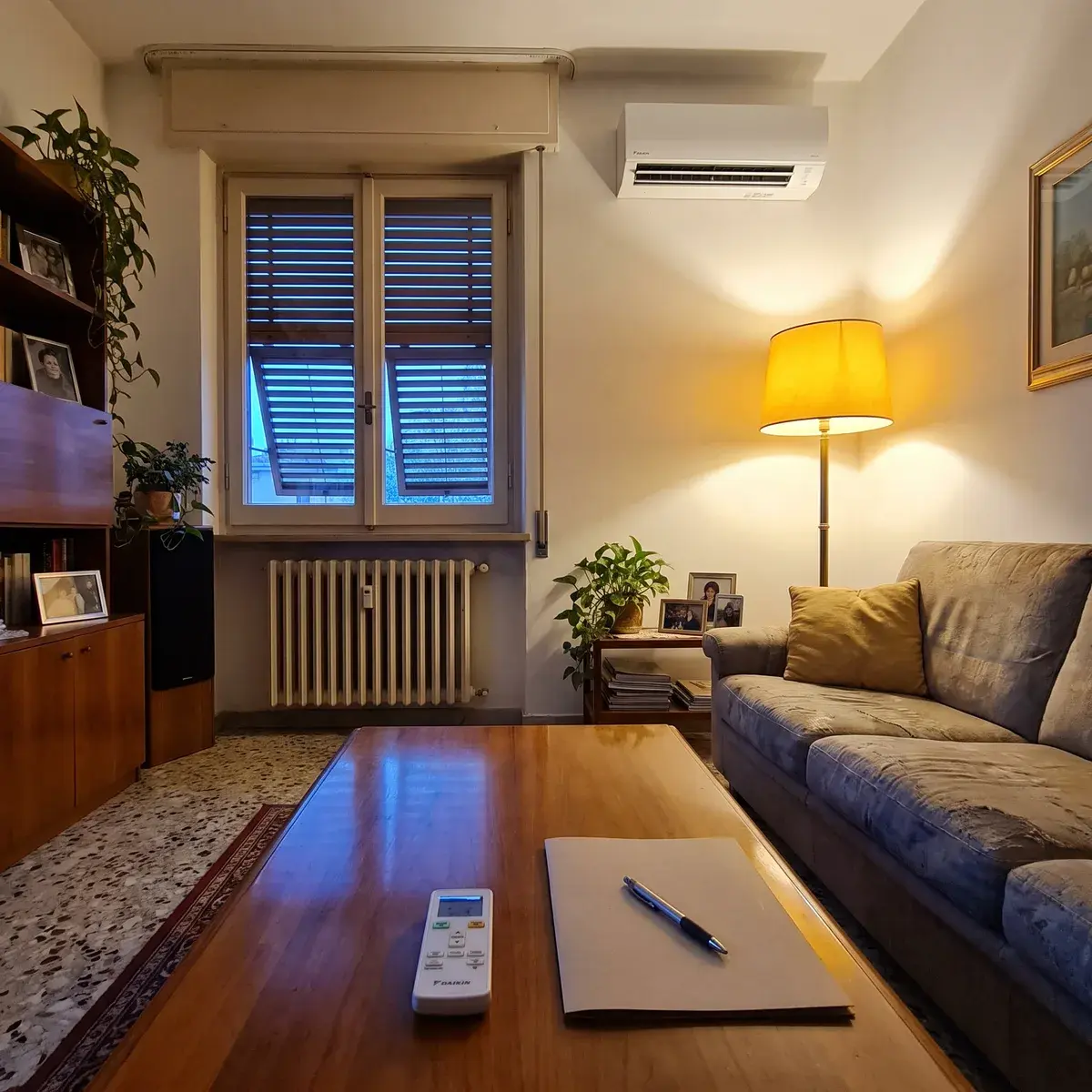 Soggiorno italiano in una sera d'inverno con un climatizzatore Daikin Perfera a parete; sul tavolino in primo piano il telecomando e una cartellina chiusa con una penna.