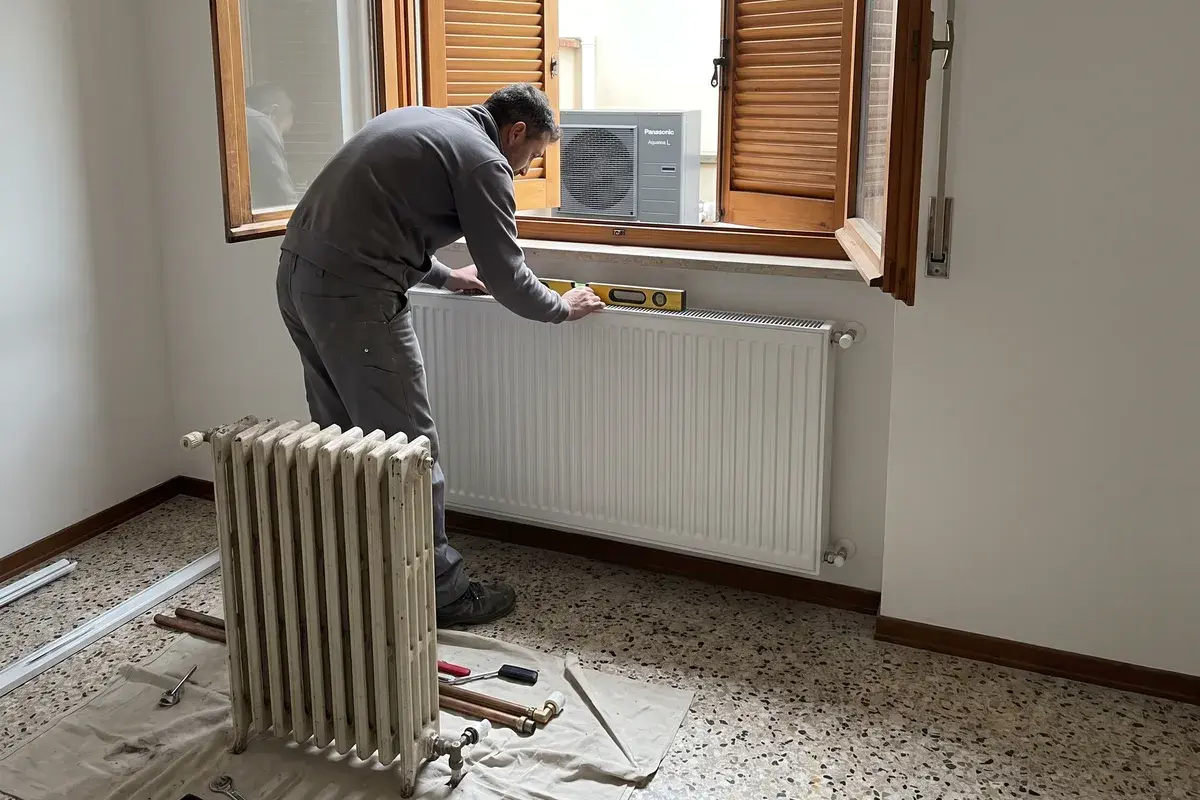Idraulico che sostituisce un vecchio radiatore in ghisa con un pannello più grande, con la pompa di calore Panasonic Aquarea L visibile dalla finestra.